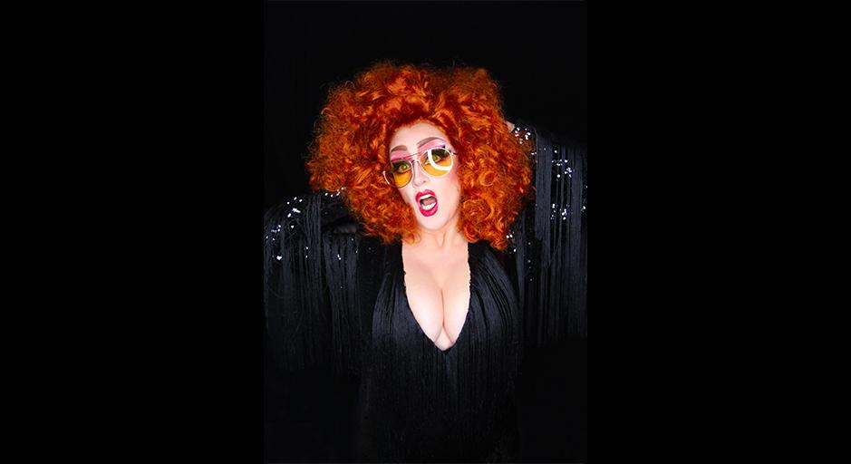 A woman with curly red hair, large amber glasses and bright red lips, in a low cut black dress
