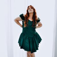 A woman in a green dress with long wavy auburn hair stands with hands on hips
