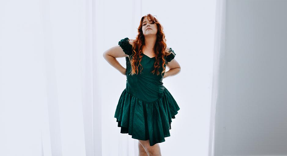 A woman in a green dress with long wavy auburn hair stands with hands on hips