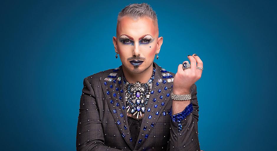 A performer with a jewelled suit, and dramatic makeup stares at the camera. 