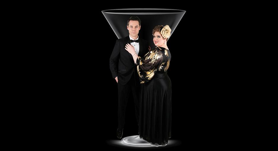 A man and a woman in black tie attire stand in front of a martini glass. 