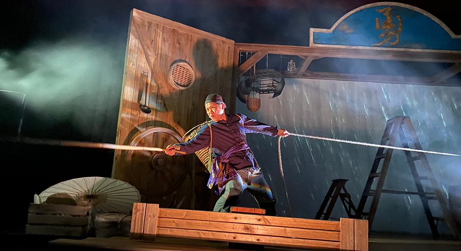 A masculine person in traditional Chinese clothing holds two ropes together in thick stage fog.