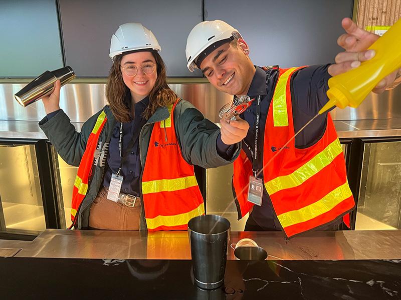 Two bar staff in high vis and hard hats make a cocktail.