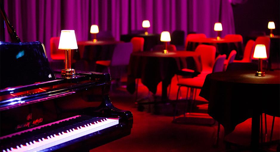 Underground caberet setting with piano in foreground, tables in background. 