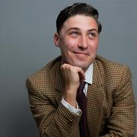 A man in a brown suit jacket smiles and looks to the right.