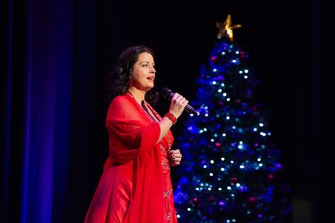 A woman in a red dress singing with a blue Christmas tree in the background. 