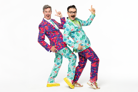 Two men in brightly coloured suits point their index fingers in the air and smile. 