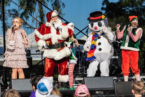 Santa and a festive snowman singing carols on stage to a crowd of children 