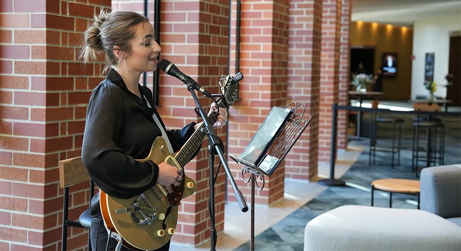 A female in a black top with her hair tied up, sings and plays guitar in The Round bar. 