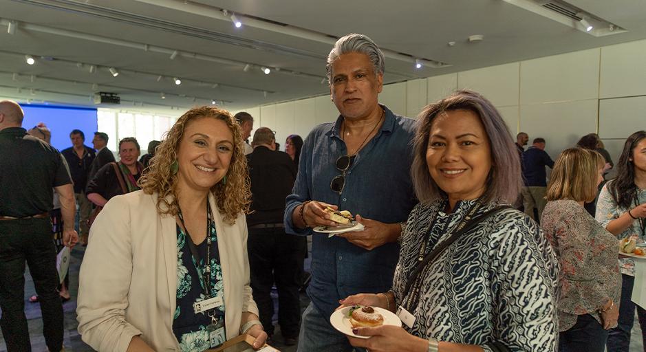 The people face the camera, each with a plate of food, with a crowd in the background inside a function room.
