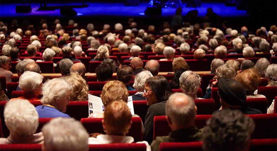 A crowd of people sit in a theatre, waiting for a show to start. 