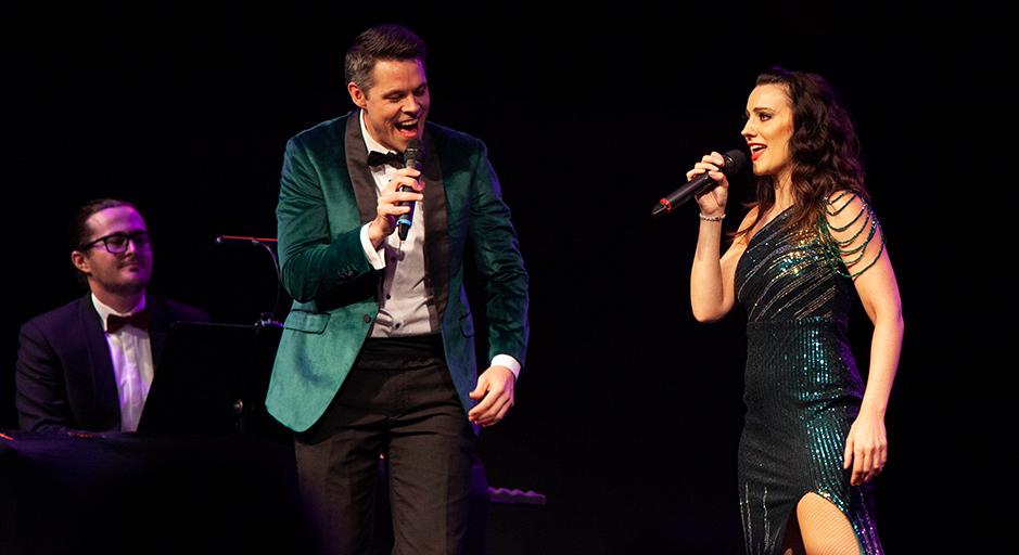 Two people sing on stage with a man on piano in the background