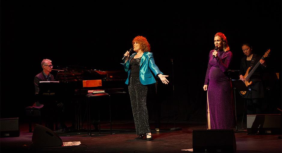 Two woman sing on stage, with a man on piano and a woman on guitar in the background 
