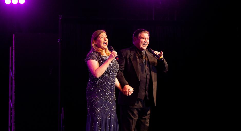 Two singers on stage, holding hands, one woman in a sparkly dress, a man in a black suit