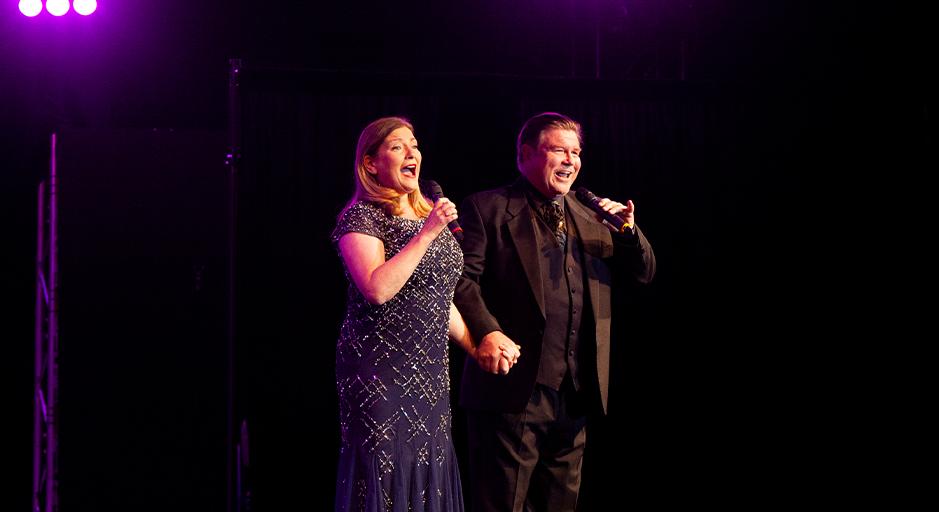 Two singers on stage, holding hands, one woman in a sparkly dress, a man in a black suit