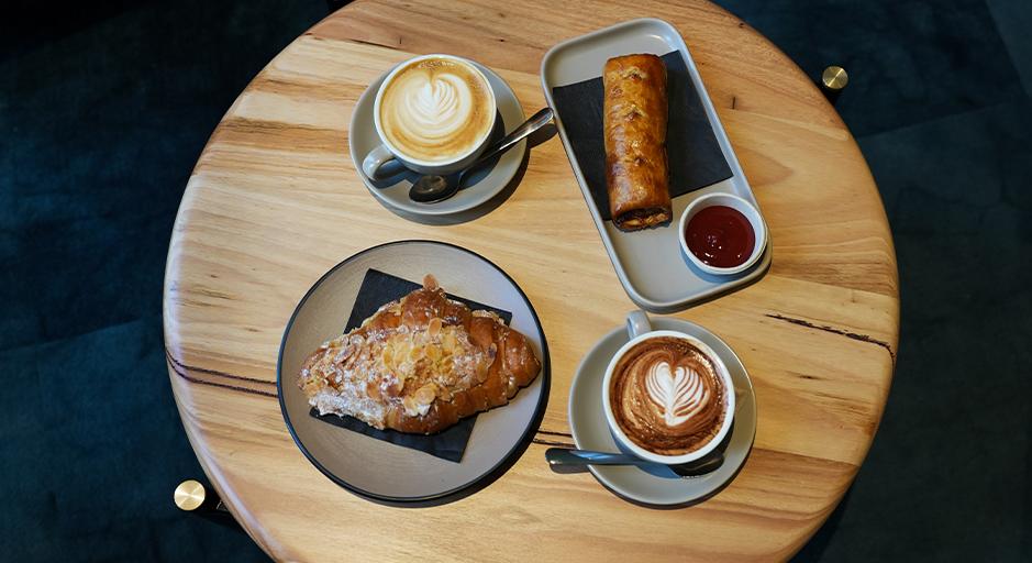 Two coffees, a sausage roll and croissant sit on a table. 