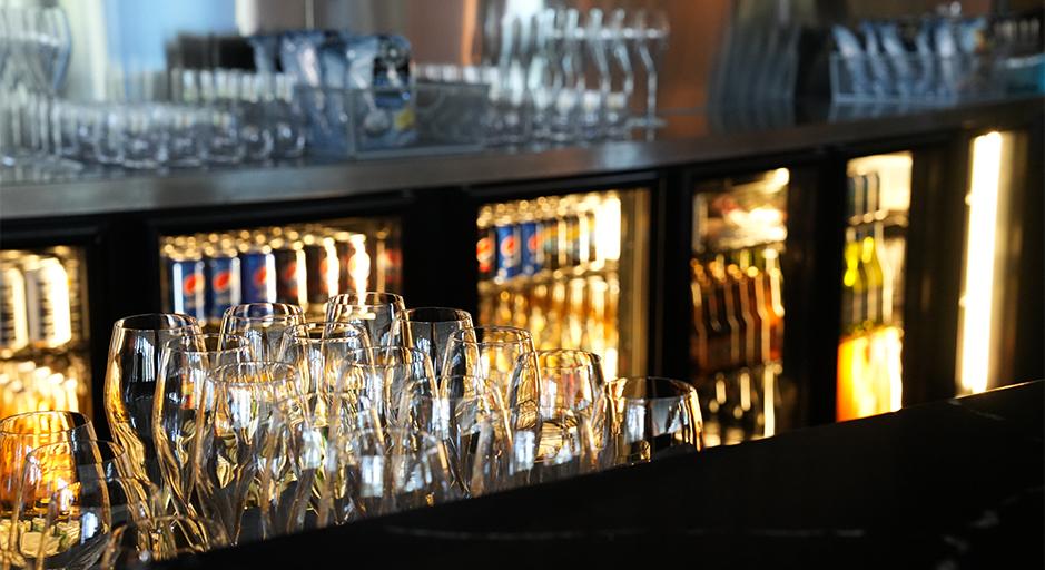 Glasses on a bar and bottles in a drinks fridge in the background