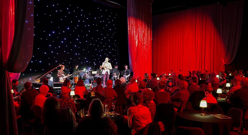 A performer sings on stage in front of an audience sitting at cabaret tables. 