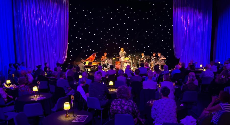 A performer sings on stage in front of an audience sitting at cabaret tables. 