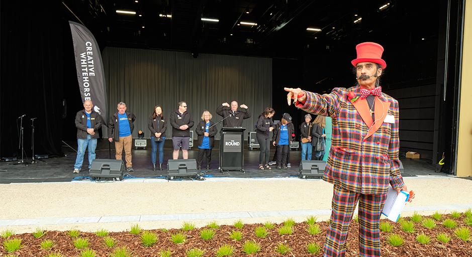 A performer in a check suit and top hat stands in front of the soundshell. 