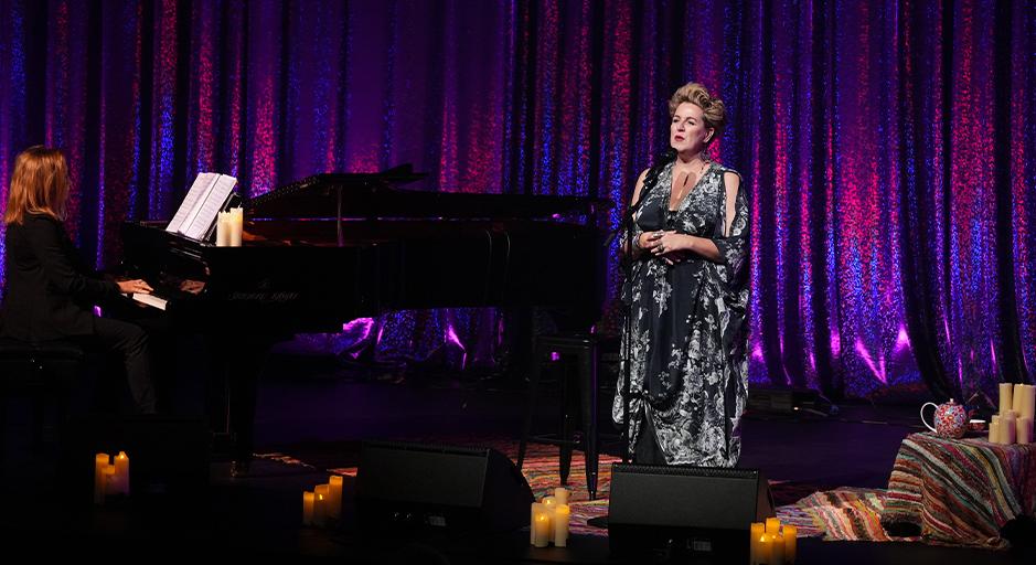 Singer Queenie van de Zandt performs on stage accompanied by a woman on piano. 